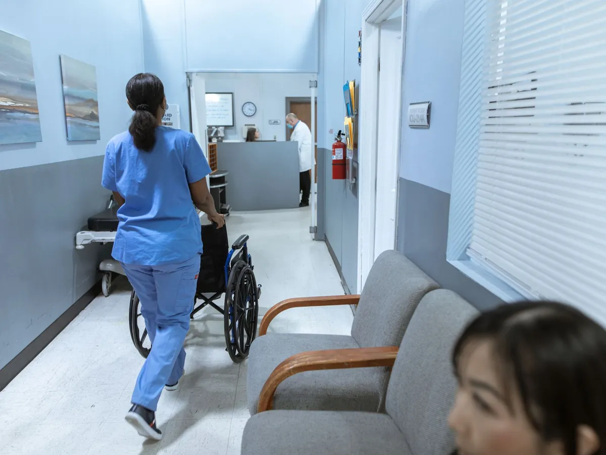 Infirmière en tenue de soins poussant un fauteuil roulant dans un couloir d'hôpital bien éclairé