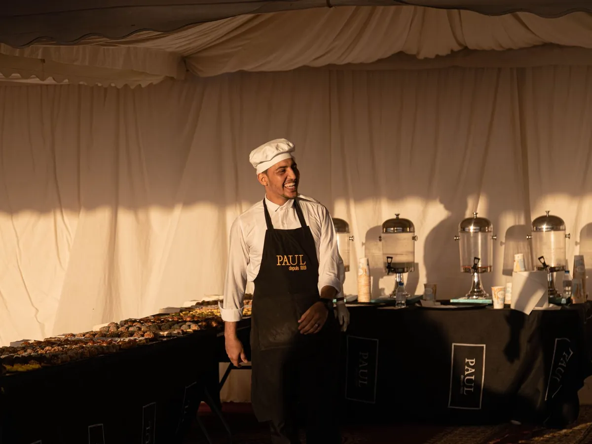 Chef souriant en tablier devant un buffet de pâtisseries, dans une ambiance accueillante