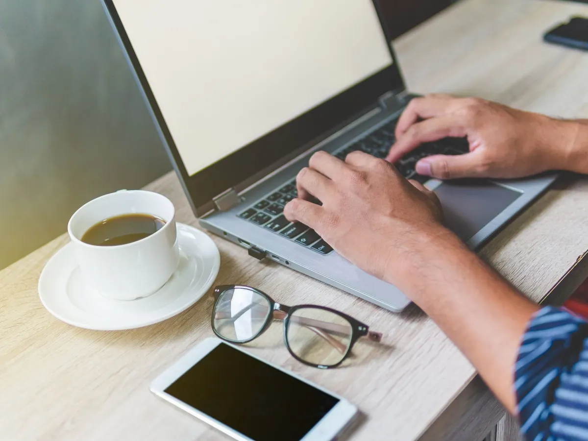 Homme tapant sur son ordinateur portable, café, téléphone et lunettes posés sur le bureau