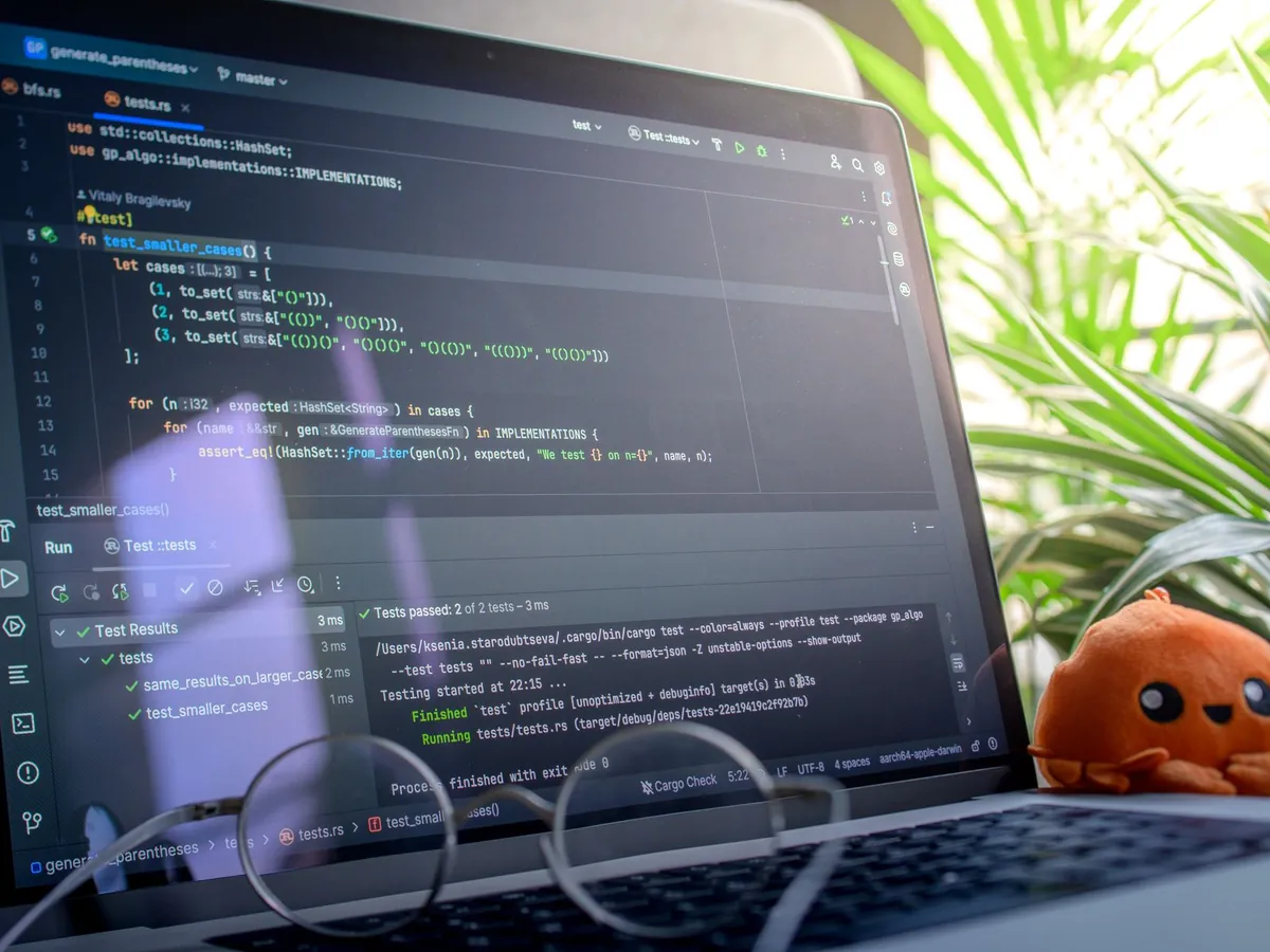 Ordinateur portable ouvert affichant du code à côté d'une peluche, dans une pièce lumineuse avec plantes