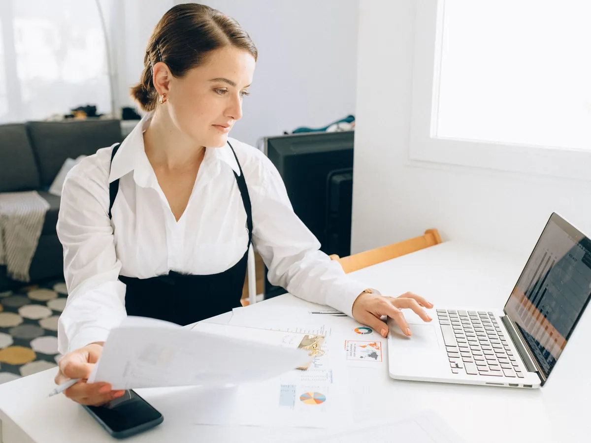 Femme à son bureau consultant des rapports d'activité sur ordinateur portable, dossiers en main