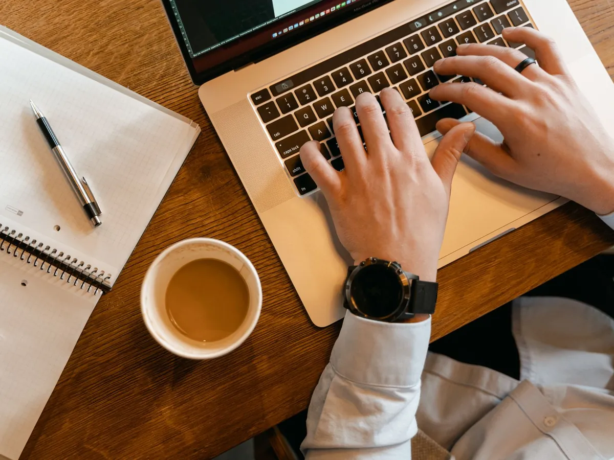 Mains tapant sur un ordinateur portable, à côté d'une tasse de café et d'un carnet sur une table en bois