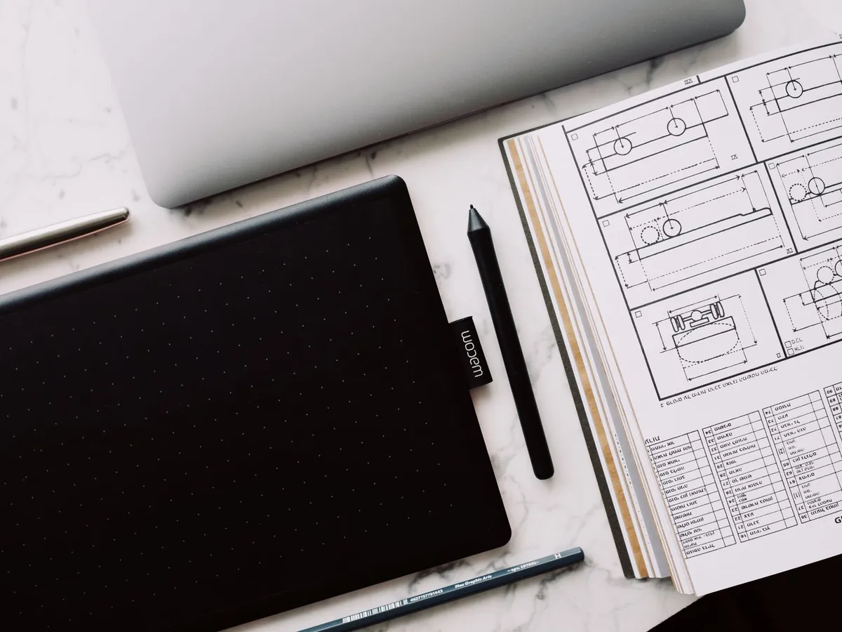 Tablette graphique noire, stylet et crayon rangés sur une table en marbre, près d'un livre ouvert