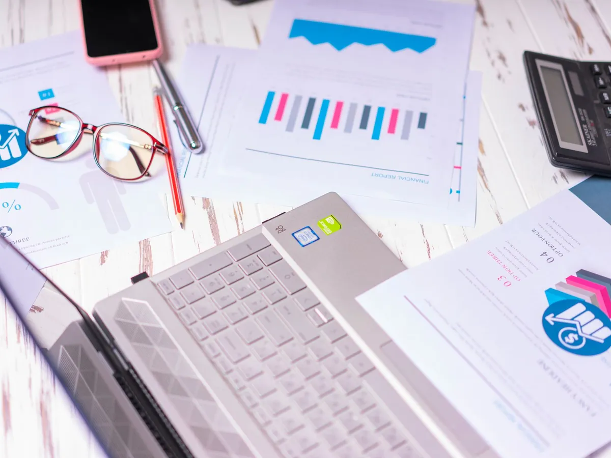 Bureau encombré : ordinateur portable, rapports financiers, calculatrice et lunettes