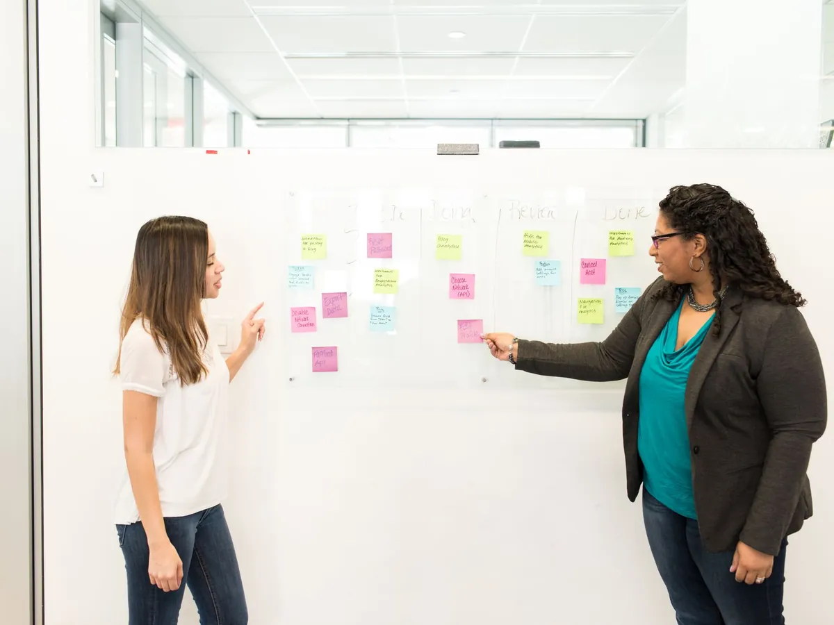 Deux femmes discutant dans un bureau, post-it sur un tableau blanc, illustrant le travail d'équipe