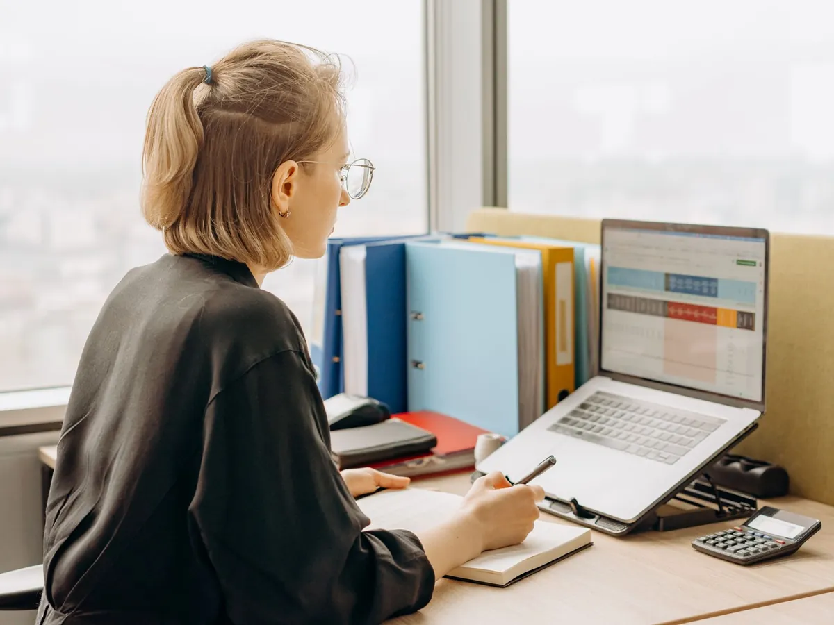 Collaboratrice à son poste de travail avec ordinateur portable, classeurs et calculatrice