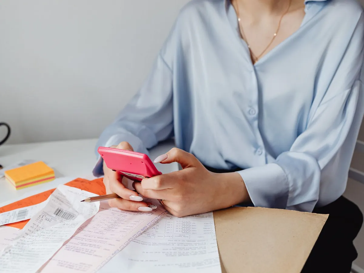 Femme utilisant une calculatrice rose, entourée de factures et de tickets de caisse sur son bureau