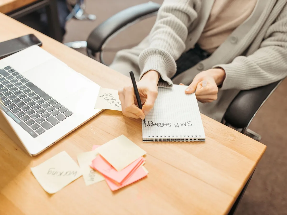 Adulte prenant des notes dans un carnet à son bureau, à côté d'un ordinateur portable