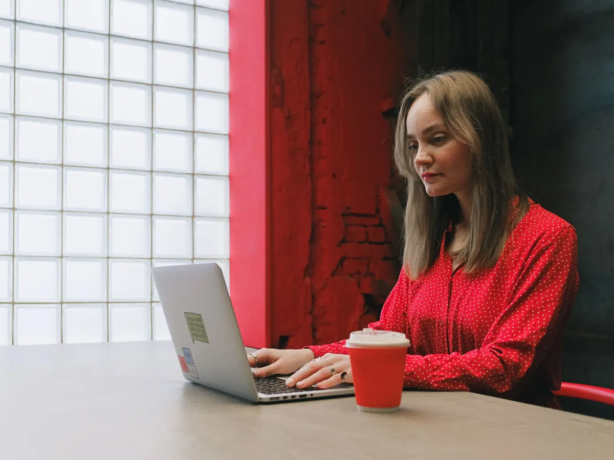 Femme en chemisier rouge concentrée sur son travail, avec ordinateur portable et café en intérieur