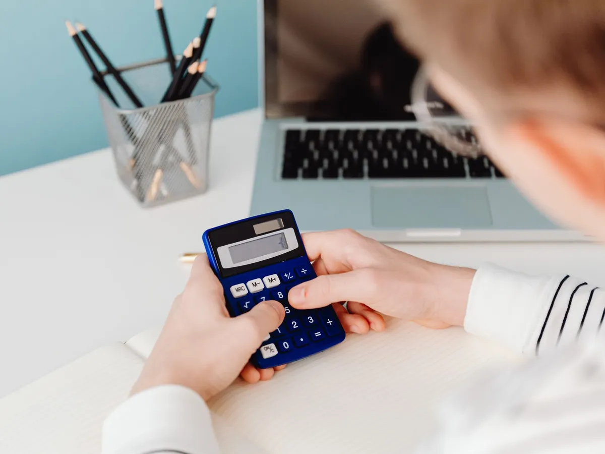 Gros plan de mains utilisant une calculatrice bleue à un bureau, avec ordinateur portable et papeterie