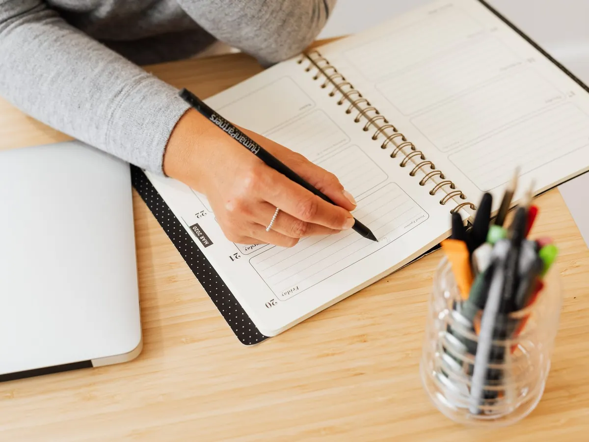 Main notant ses priorités dans un agenda papier à côté d'un ordinateur portable