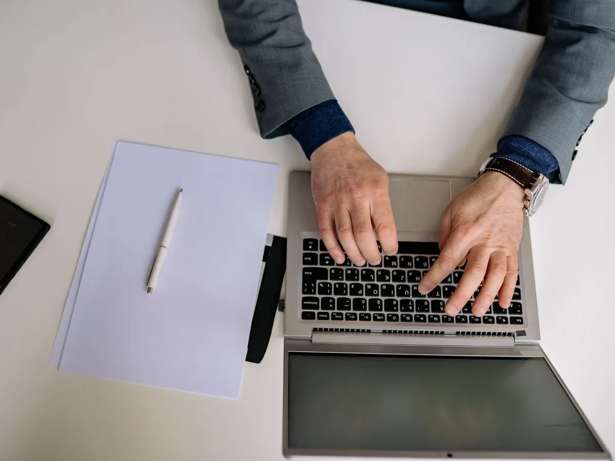 Vue de dessus d'un homme en costume tapant sur son ordinateur portable, bloc-notes et stylo à proximité