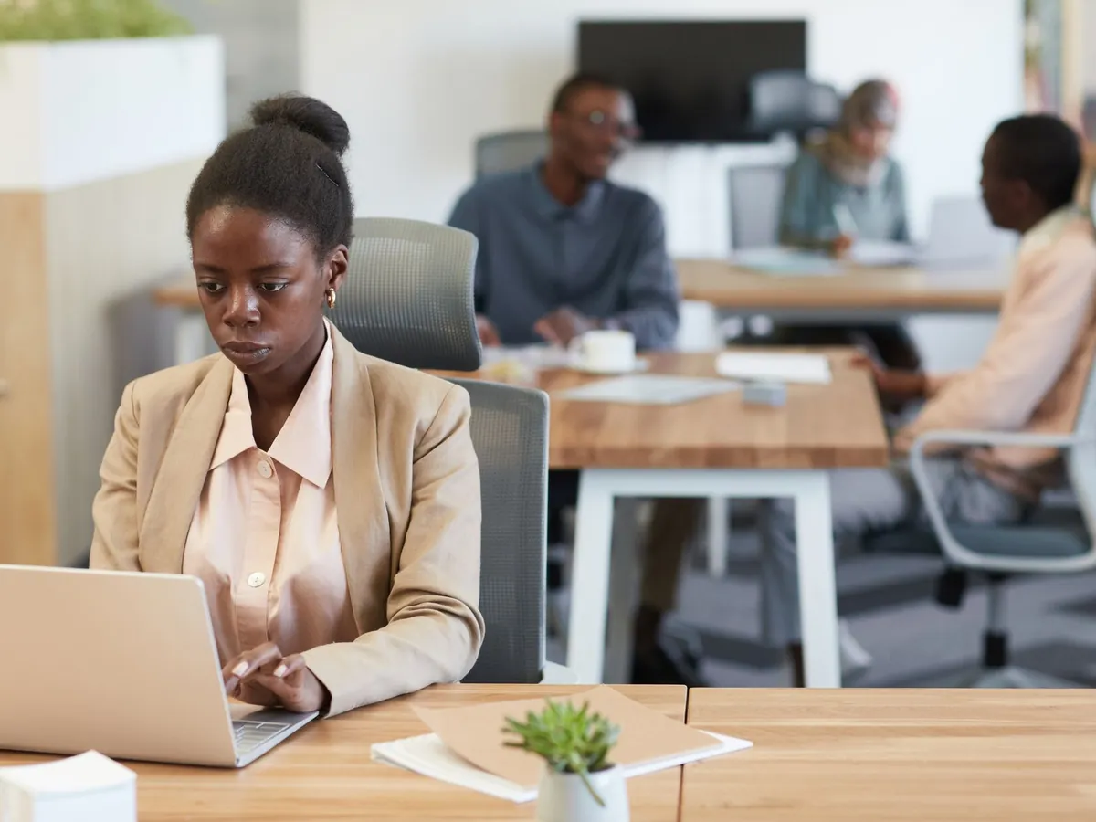 Jeune femme africaine en blazer, concentrée sur son ordinateur portable dans un bureau animé