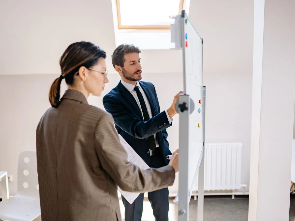 Deux collègues en tenue formelle discutant de stratégies sur un tableau blanc dans un espace moderne