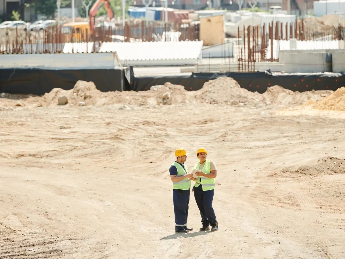 Deux ouvriers discutant de plans sur un chantier sablonneux, en équipement de sécurité