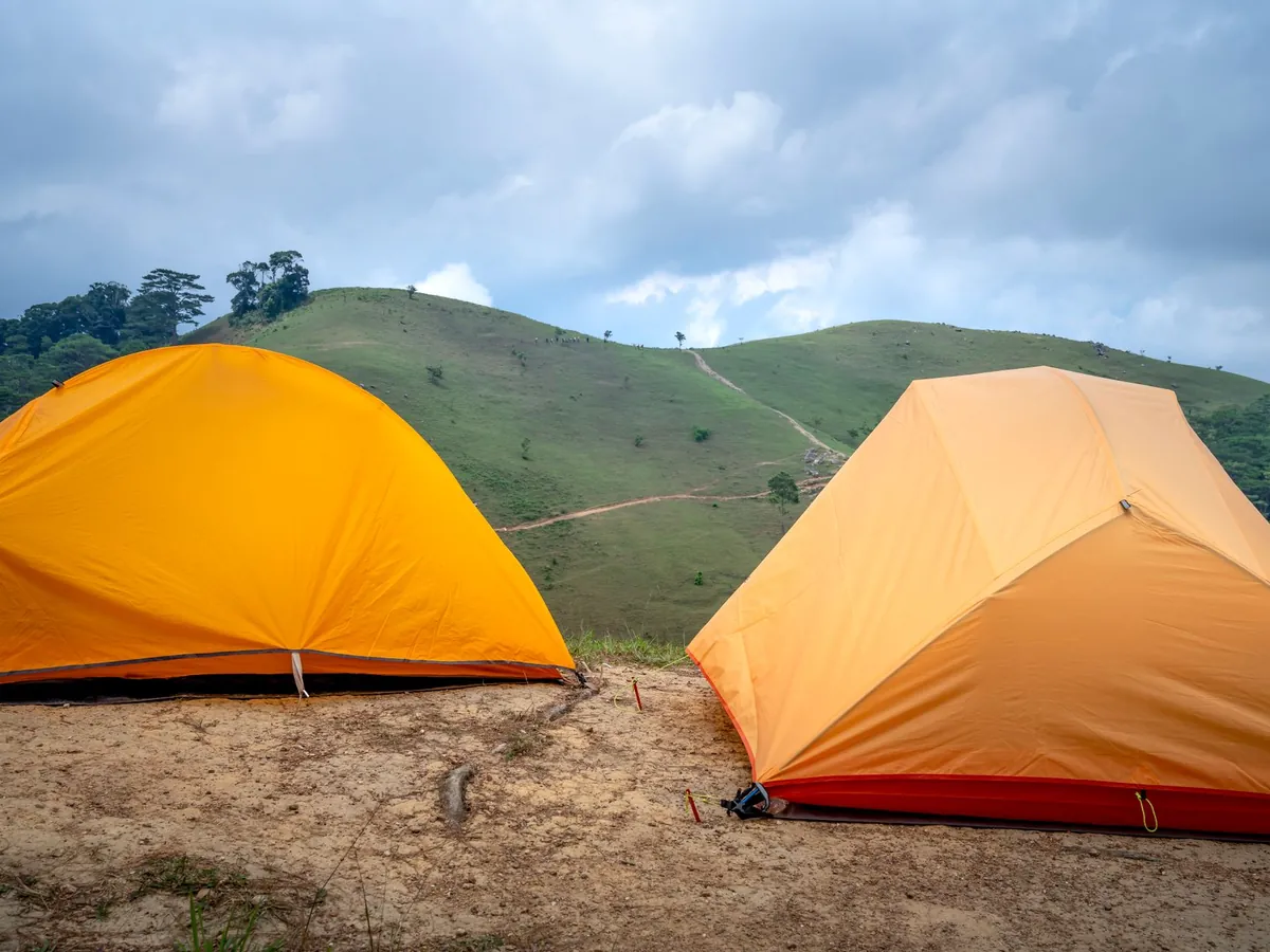 Tentes de camping orange sur un sol sablonneux, au pied d'une montagne verte sous un ciel nuageux d'été