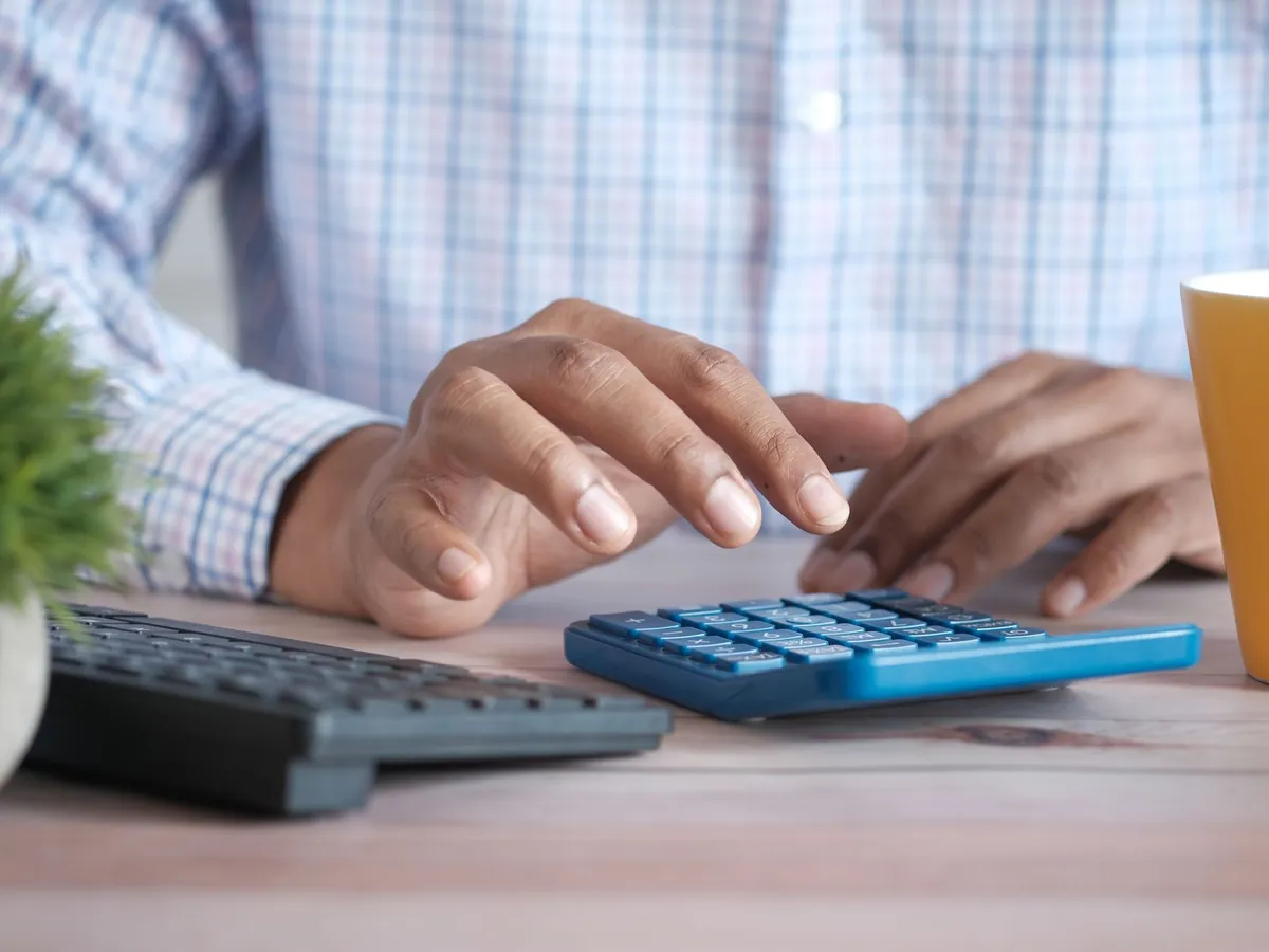 Personne utilisant une calculatrice bleue sur un bureau en bois, avec un clavier et une plante à proximité