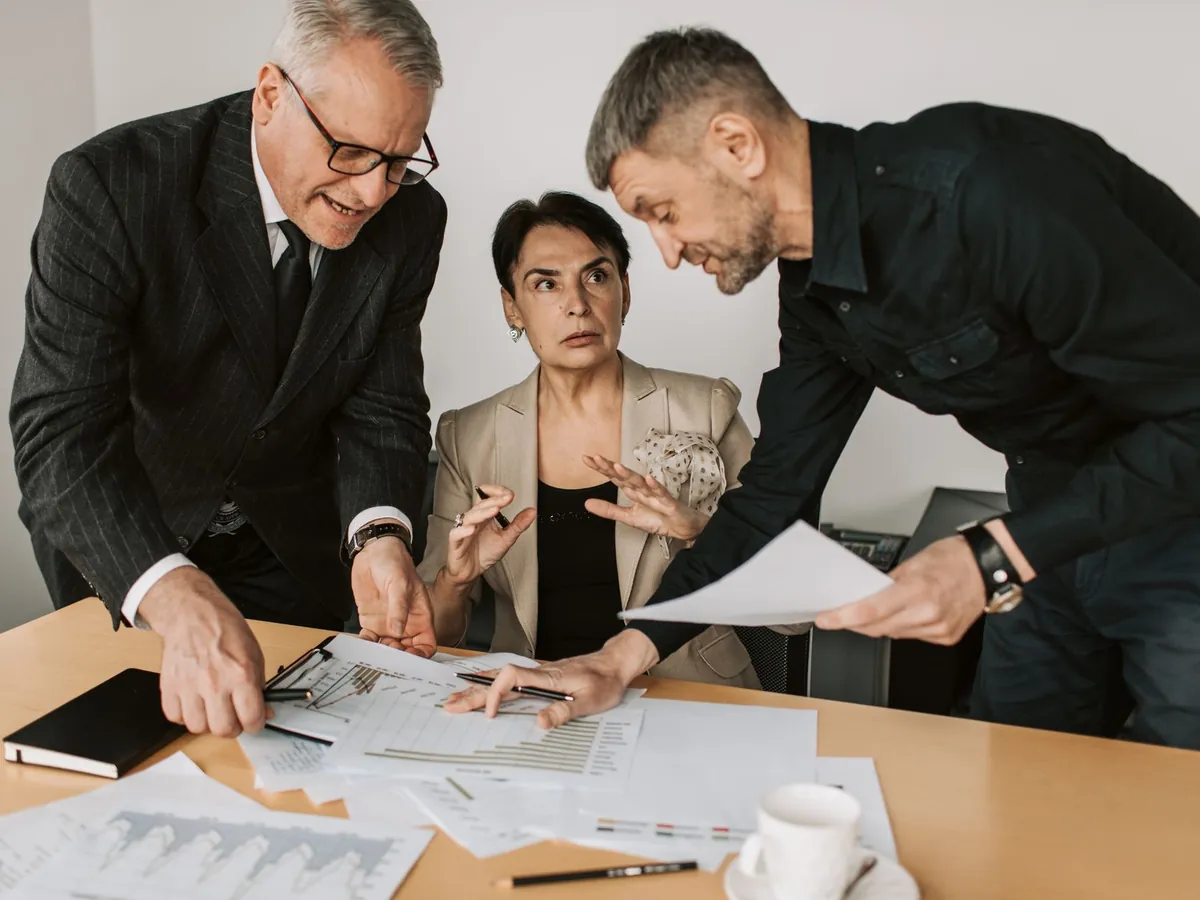Trois adultes en discussion dynamique autour d'une table de bureau, analysant des documents
