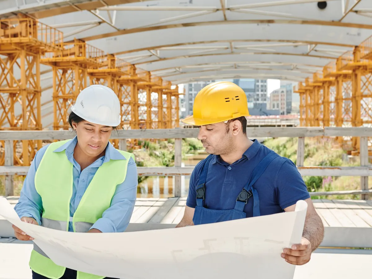Ingénieure et chef de chantier en casque examinant des plans sous une structure métallique